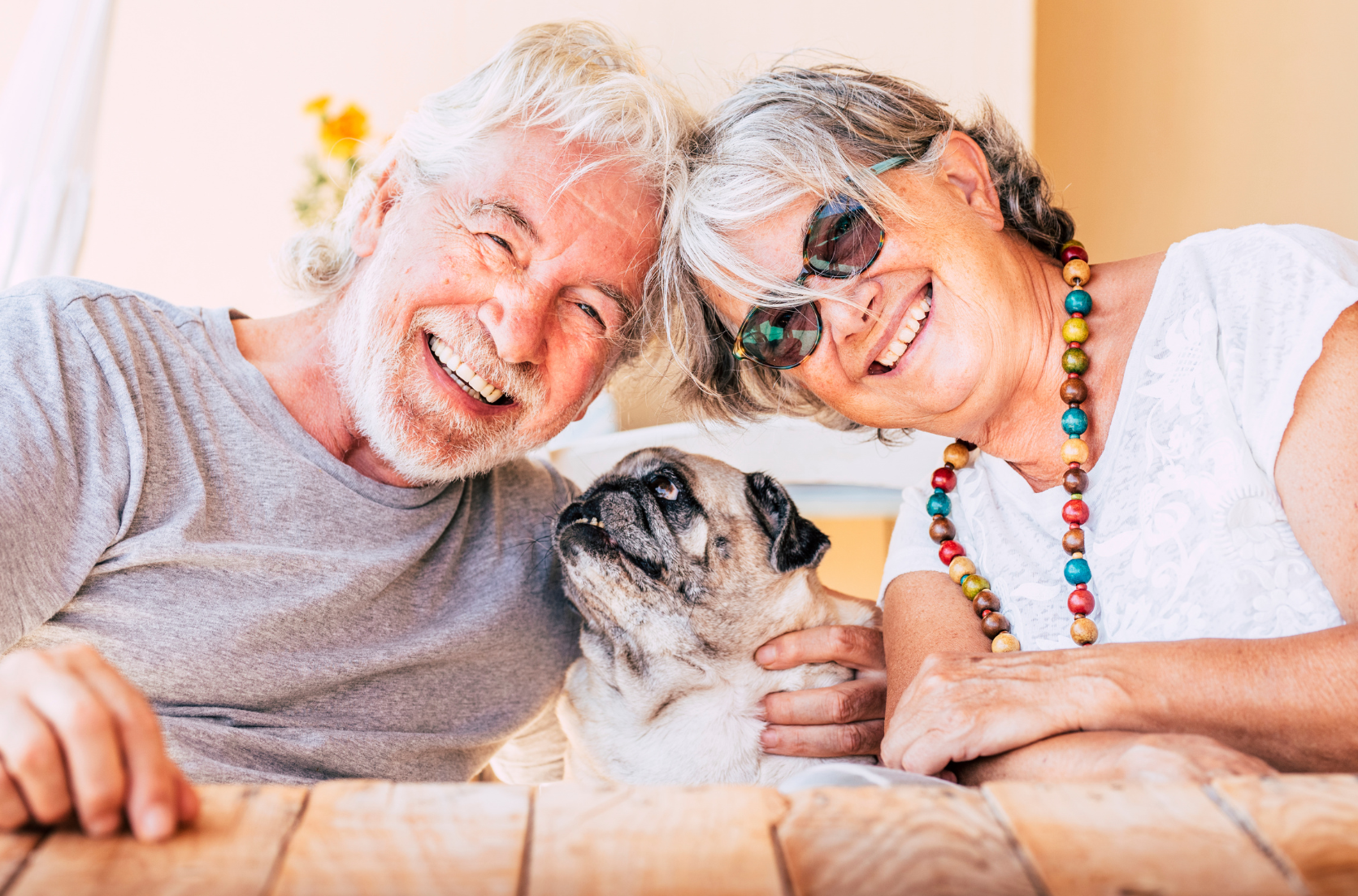 elderly couple Smiling at the camera with their head together. Small pug is in the middle of them looking up at the man smiling. 