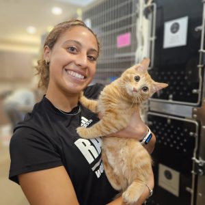 Young woman in a black shirt smiling holding an orange tabby cat looking at the camera.