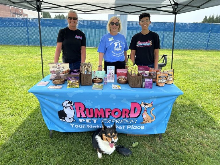 Summertime event at URI soccer fields with Rumford pets. Small black, white and tan corgi laying under the table. Three women lined up behind the table.