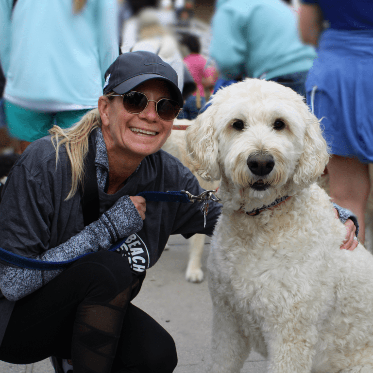 woman knelt down to pose with her white golden doodle, they are both smiling.